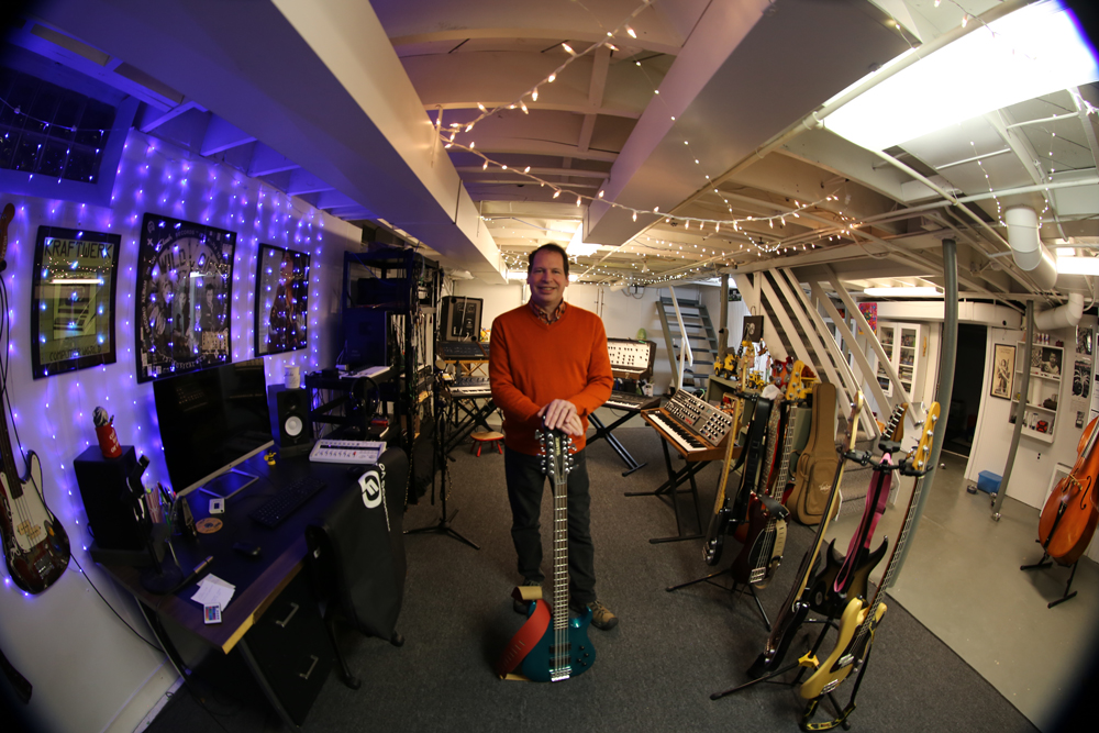 Person standing with a guitar, surrounded by musical instruments in a room with twinkling lights strung across the ceiling and hanging down the walls.
