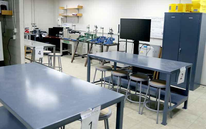 lab room with round stools underneath three tables, computer monitor screen, other lab equipments set on top of tables by the wall